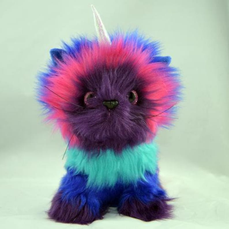 Op de afbeelding: Een pluche kat met een eenhoornhoorn. De kat is een mix van kleuren, waaronder paars, roze, blauw en turquoise. Het heeft een pluizig uiterlijk en glinsterende ogen. Het speelgoed is ontworpen om rechtop te zitten.