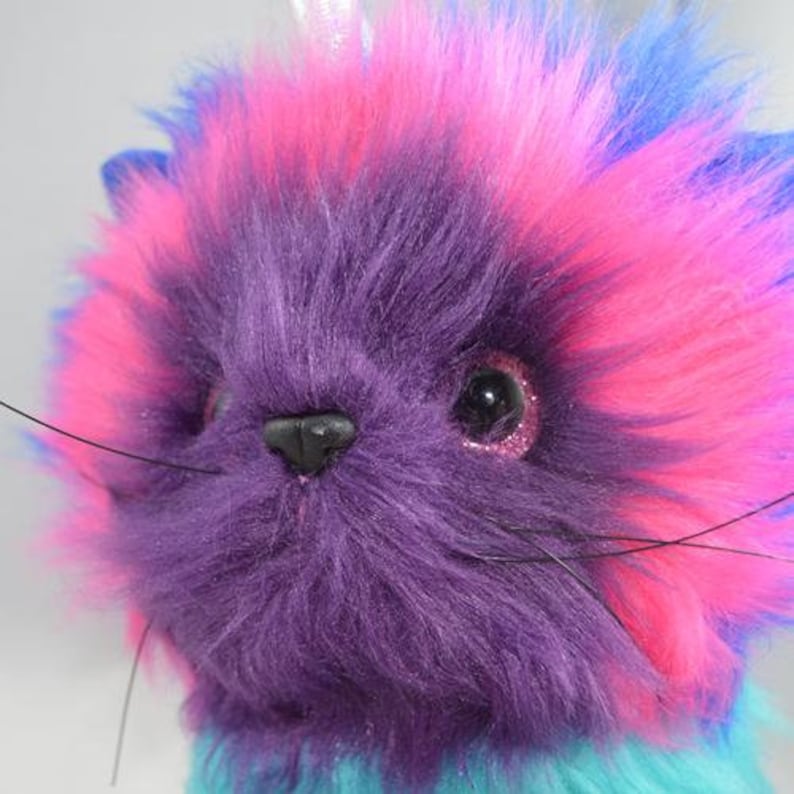 Op de afbeelding: Een close-up van een pluche kat met levendige kleuren. De vacht van de kat is een mix van paars, roze en blauw, met glinsterende ogen en lange snorharen. Het pluche speelgoed heeft een zachte, donzige textuur en een zwarte neus. Het thema is een grillige, kleurrijke kat.