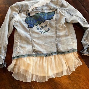 Könnte beinhalten: Hellblaue Jeansjacke mit weißem Rüschensaum und einem dekorativen Aufnäher auf dem Rücken. Der Aufnäher zeigt ein blaues und grünes Design mit den Worten "Slow Train". Über dem Aufnäher befindet sich ein weißer, federartiger Akzent.