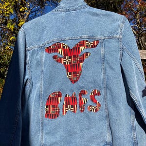 Könnte beinhalten: Hellblaue Jeansjacke mit einem dekorativen Aufnäher mit einem rot-schwarz-weißen geometrischen Muster in Form eines Bullenkopfes und dem Wort "OWES". Die Jacke hängt draußen.