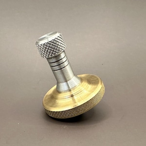 May include: A metal spinning top with a brass base and a silver-colored top. The base has a textured, ridged design, and the top has a knurled grip. The top is angled slightly, set against a neutral background.