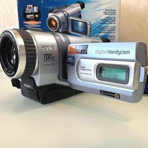 May include: A silver Sony Digital8 Handycam camcorder with a 700x digital zoom and 25x optical zoom lens. The device features a flip-out LCD screen displaying a digital timer, Super SteadyShot technology, and USB streaming capabilities.