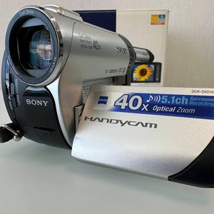 Op de afbeelding: Een zilverkleurige Sony Handycam DCR-DVD109 camcorder met een Carl Zeiss Vario-Tessar lens. Het apparaat beschikt over 40x optische zoom, 5.1-kanaals surround-opname en een uitklapbaar scherm. De camerabehuizing is zilver en zwart met merknaam.