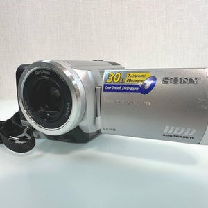 Puede incluir: Una videocámara Sony Handycam DCR-SR40 plateada con lente Carl Zeiss Vario-Tessar. El dispositivo cuenta con un disco duro de 30GB, capacidad One Touch DVD Burn y una correa de muñeca negra. El texto en el cuerpo incluye 30GB, 7hrs High Quality, 20hrs Long Play y HDD Hard Disk Drive.