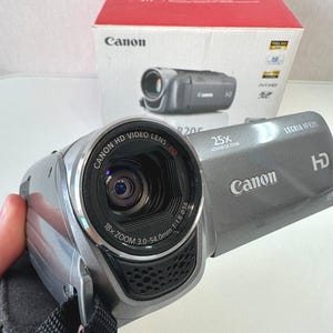 Op de afbeelding: Een zilverkleurige Canon Legria HF R205 HD-camcorder met 25x geavanceerde zoom, 18x optische zoom en een brandpuntsafstand van 3,0-54,0 mm. Het apparaat heeft een zwarte handriem en staat voor de originele wit-rode productdoos.