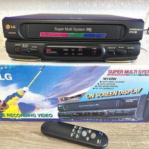 Könnte beinhalten: Ein schwarzer LG W42W Super Multi System VHS-Videorekorder mit Frontlader-Kassettenschacht, Bedientasten und Digitalanzeige. Das Gerät wird mit einer schwarzen Fernbedienung und der originalen Pappverpackung geliefert.