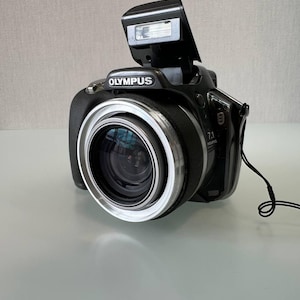Peut inclure: Un appareil photo numérique Olympus noir doté d'un objectif proéminent avec un anneau argenté, d'un flash escamotable et d'une étiquette 7.1 mégapixels sur le côté. L'appareil comprend une dragonne noire fixée sur le côté et une poignée texturée.