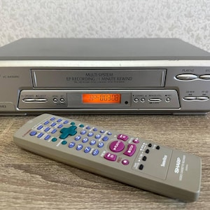 Puede incluir: Un reproductor de video VHS Sharp VC-AA350RU plateado con pantalla multisistema, grabación EP y función de rebobinado de 1 minuto. El dispositivo incluye un control remoto gris a juego con botones morados y un pad direccional verde.