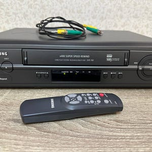 Peut inclure: Un lecteur VHS Samsung gris foncé avec une fente de chargement frontale, marqué x400 SUPER SPEED REWIND, LONG PLAY SYSTEM et SVR-160. Il comprend une télécommande gris foncé assortie et un câble noir avec des connecteurs RCA jaune et vert.