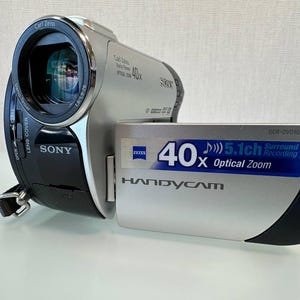 May include: A silver and black Sony Handycam DCR-DVD109 camcorder with a Carl Zeiss Vario-Tessar lens. The side panel features text reading 40x Optical Zoom and 5.1ch Surround Recording. The device includes a lens cover switch and a strap attachment.