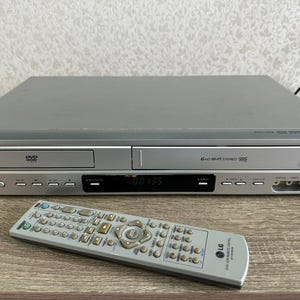 Peut inclure: Un lecteur combiné DVD et VHS LG de couleur argentée avec un tiroir à disque à gauche et une fente pour cassette VHS à droite. La façade comprend un écran numérique, des boutons de commande et des ports d'entrée. Une télécommande argentée est posée devant.