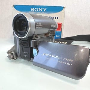 Puede incluir: Una videocámara digital Sony Handycam gris con lente Carl Zeiss Vario-Sonnar, pantalla LCD panorámica abatible y marca de 3.0 megapíxeles. El dispositivo cuenta con una correa negra y está colocado frente a su caja original azul y blanca.