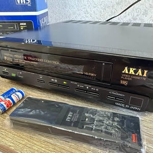 Może przedstawiać: Czarny odtwarzacz kaset wideo VHS Akai z pilotem i bateriami. Odtwarzacz ma napis "Digital Remote Tracking Control" i "Made in Japan". Pilot jest w plastikowym opakowaniu. Odtwarzacz znajduje się na jasnej powierzchni.