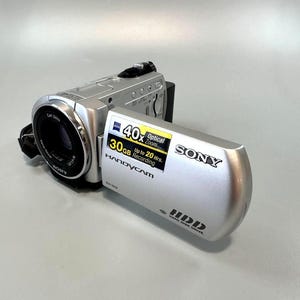 May include: A silver Sony Handycam camcorder with a Carl Zeiss Vario-Tessar lens. The open side panel displays text including 40x Optical Zoom, 30GB Up to 20 Hrs. Recording, Sony, Handycam, and HDD Hard Disk Drive. Model number DCR-SR32 is visible.