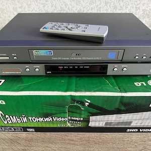 Peut inclure: Un lecteur de cassettes vidéo VHS LG gris avec une télécommande assortie posée dessus. L'appareil dispose d'une fente de chargement frontale, de boutons de commande et d'un écran numérique. Il est posé sur sa boîte en carton verte et blanche.