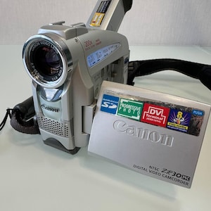 Può includere: Una videocamera digitale Canon ZR30MC color argento con schermo LCD ribaltabile. Il dispositivo è dotato di un obiettivo zoom digitale 200x, terminale DV e compatibilità con schede SD. Lo schermo mostra etichette per Progressive Photo e 8 ore di registrazione.