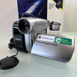 Może przedstawiać: Srebrno-czarna kamera Sony Handycam DCR-HC27E z obiektywem Carl Zeiss Vario-Tessar, 20-krotnym zoomem optycznym i dotykowym ekranem LCD. Urządzenie znajduje się przed oryginalnym niebiesko-białym kartonowym opakowaniem.