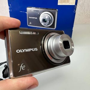 May include: A brown Olympus FE digital camera with a silver lens and the word "OLYMPUS" printed on the front. The camera is held in a hand, with a blue box in the background that also displays the camera.