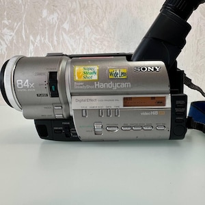 Peut inclure: Caméscope Sony Handycam argenté avec un objectif noir. L'appareil photo porte les inscriptions "84x Digital Zoom", "Wide Lens" et "Super SteadyShot". Il possède un écran numérique et divers boutons.