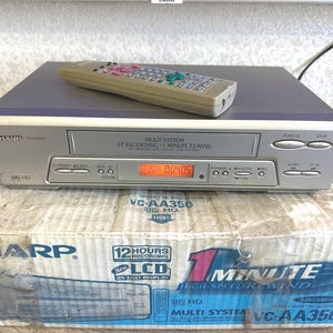 Peut inclure: Un magnétoscope VHS Sharp VC-AA350 argenté avec un écran numérique affichant 00405, doté d'une étiquette multi-système et d'une télécommande posée dessus. L'appareil est placé sur son emballage en carton d'origine.