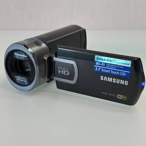 May include: A black Samsung HMX-QF20 camcorder with a Schneider Kreuznach Varioplan lens. The device features a 2.7-inch smart touch LCD screen, Full HD recording capabilities, Wi-Fi connectivity, and Switch Grip II technology for vertical recording.