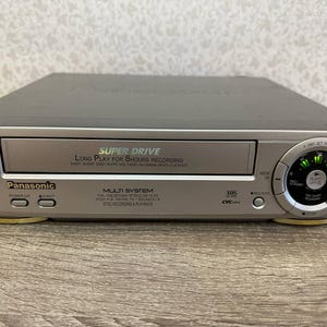 Puede incluir: Videograbadora Panasonic SJ5 plateada con texto "Super Drive" y "Long Play for 8 hours recording". Cuenta con un diseño multisistema y compatibilidad VHS. Incluye botones de encendido y expulsión, y un panel de control circular.