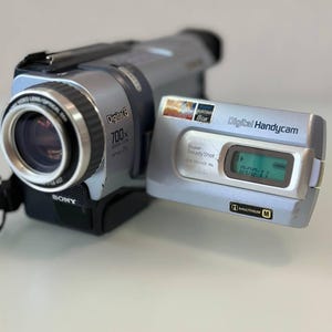 May include: A silver Sony Digital8 Handycam camcorder with a flip-out screen displaying text. The device features a 700x digital zoom lens, Super SteadyShot technology, and an InfoLithium M battery indicator. The model number DCR-TRV240E PAL is visible.