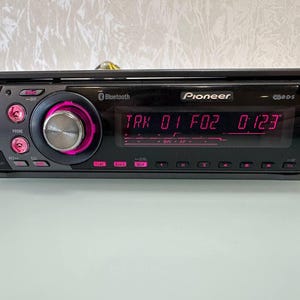 May include: A black Pioneer car stereo with a digital display showing "TRK 01 F02 0123". The stereo has pink illuminated buttons and a silver control knob. Features Bluetooth connectivity and CD-R/S compatibility.