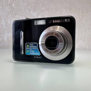 May include: Black Samsung D860 digital camera with a silver lens and flash. Features include face detection and digital image stabilization. The lens has a zoom range of 6.3-18.9mm.