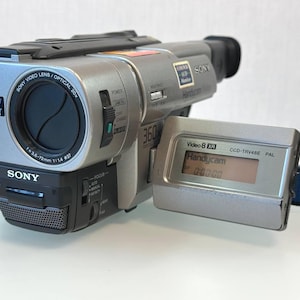 May include: A silver and black Sony Handycam camcorder with a flip-out LCD screen displaying the text Handycam 0:00:00. The lens barrel features text including Sony Video Lens Optical 20x, NightShot 0 Lux, and f=3.6-72mm 1:1.4 37.