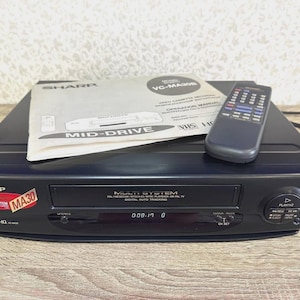 May include: A black Sharp VC-MA30B multi-system VHS video cassette recorder featuring a front-loading tape slot, digital auto tracking, and a digital display. Included are a matching black remote control and an operation manual with the text MID-DRIVE.