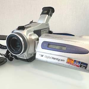 Może przedstawiać: Srebrna kamera Sony MiniDV Digital Handycam z obiektywem Carl Zeiss Vario-Sonnar, 120-krotnym zoomem cyfrowym i technologią Super SteadyShot. Urządzenie posiada rozkładany ekran LCD z napisami Digital Handycam oraz Super NightShot.
