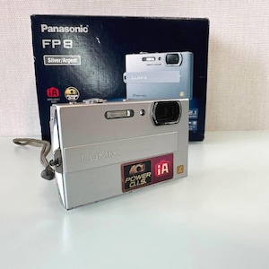 May include: A silver Panasonic Lumix FP8 digital camera with a wrist strap, featuring a rectangular body, a lens, and a flash. The camera is positioned in front of its original dark blue product box, which displays the camera model and branding.