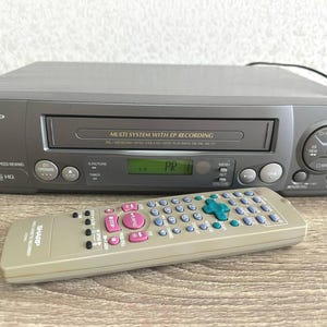 Könnte beinhalten: Ein dunkelgrauer Sharp VC-VA360 VHS-Videorekorder mit Digitalanzeige, Frontlader-Kassettenschacht und Wiedergabesteuerung, begleitet von einer passenden hellgrauen Fernbedienung mit zahlreichen Tasten und einem grünen Steuerkreuz.