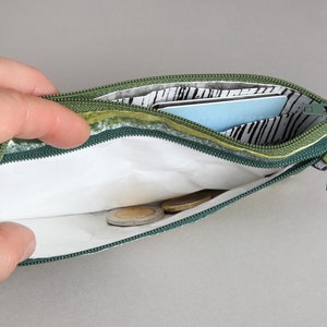 May include: A green zippered pouch with a white lining and a black and white patterned fabric. The pouch is open and contains a few coins and a blue card.