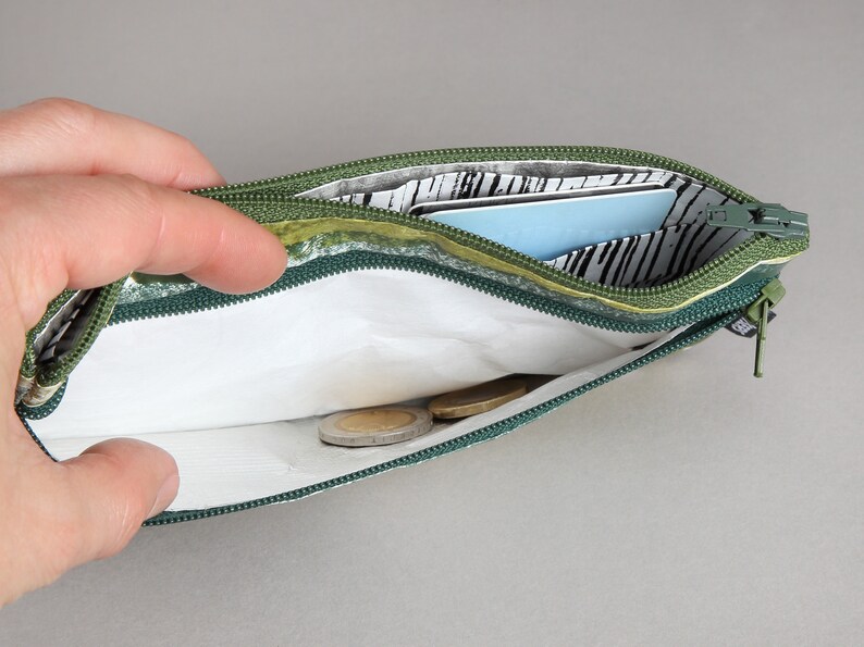 May include: A green zippered pouch with a white lining and a black and white patterned fabric. The pouch is open and contains a few coins and a blue card.