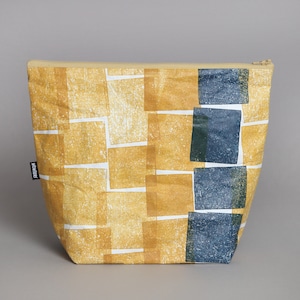 Könnte beinhalten: Eine gelbe und blaue Reißverschlusstasche mit geometrischem Muster. Die Tasche ist aus strukturiertem Stoff und hat ein schwarzes Etikett mit dem Wort "monokel" darauf.