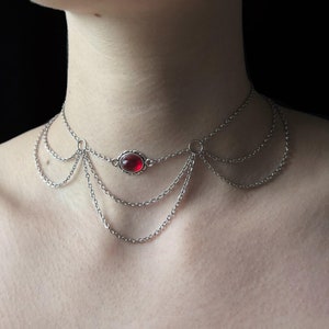 Può includere: Un girocollo a catena d'argento con un ciondolo di pietra rossa. La collana ha più strati di catene.