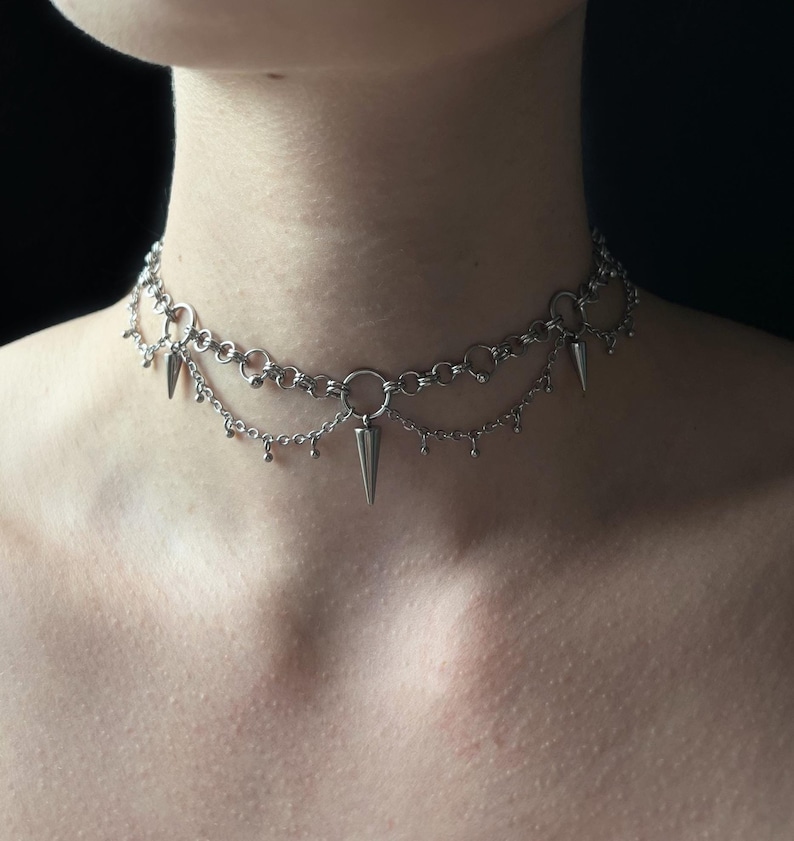 May include: A silver chain choker necklace with multiple dangling spikes. The necklace is layered with two chains, one with a larger chain link and the other with a smaller chain link. The spikes are all pointed and are attached to the larger chain link.