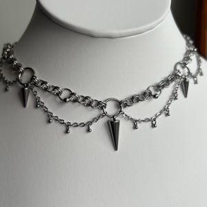 May include: A silver chain choker necklace with multiple layers and dangling spikes. The necklace is on a white mannequin bust.