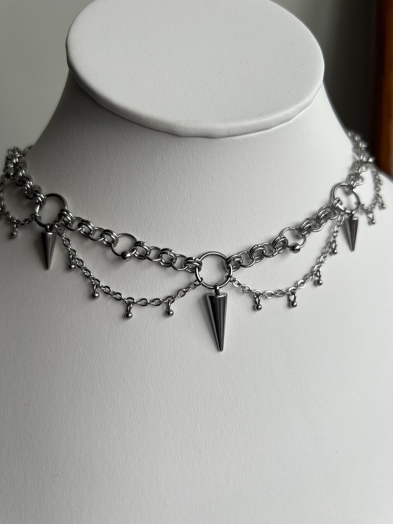 May include: A silver chain choker necklace with multiple layers and dangling spikes. The necklace is on a white mannequin bust.