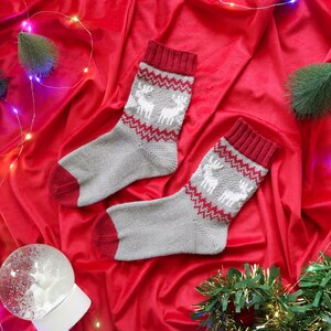 May include: Two pairs of grey and red Christmas socks with a white reindeer pattern on a red velvet background with Christmas lights and decorations.