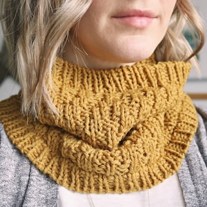 May include: A close-up of a mustard yellow knitted cowl neck scarf. The scarf is folded over and worn around the neck.