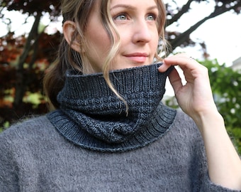 May include: A dark gray knitted cowl neck warmer. The cowl is a deep charcoal color with a ribbed texture and a folded design. The model is wearing a gray sweater.