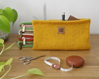 May include: A yellow knitted pouch with a brown leather label that says "SONG BIRD". The pouch is filled with knitting needles, a pen, and a small leather wallet. There are also several smaller zippered pouches stacked next to the larger pouch. The pouches are on a wooden surface with a green plant in the background.