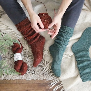 Peut inclure: Deux paires de chaussettes tricotées à la main, l'une de couleur rouille et l'autre de couleur vert sarcelle. Les chaussettes de couleur rouille sont en cours de réalisation avec une aiguille à tricoter rouge.