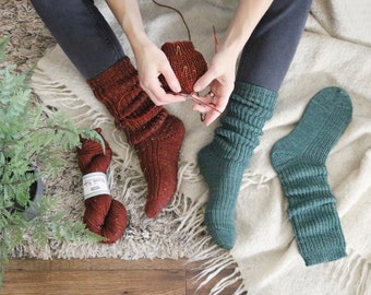 May include: Two pairs of hand-knitted socks, one in a rust color and one in a teal green. The rust colored socks are being worked on with a red knitting needle.