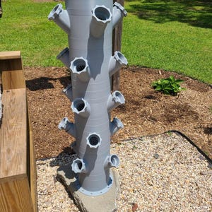 May include: A gray vertical garden planter with multiple angled openings for plants. The planter is cylindrical with several rows of openings, and is mounted on a stone base. The background includes grass, mulch, and a wooden fence.