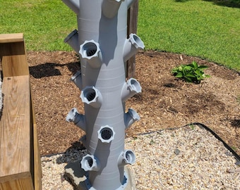 May include: A gray vertical garden planter with multiple angled openings for plants. The planter is cylindrical with several rows of openings, and is mounted on a stone base. The background includes grass, mulch, and a wooden fence.