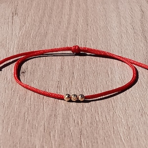 May include: A red string bracelet with three gold beads. The bracelet is tied with a knot and has a simple design.